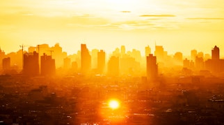 The photo depicts Sunrise over modern office buildings in business district center of Bangkok. Skyline view of cityscape with sunlight and flare in warm light color tone. Construction business concept
