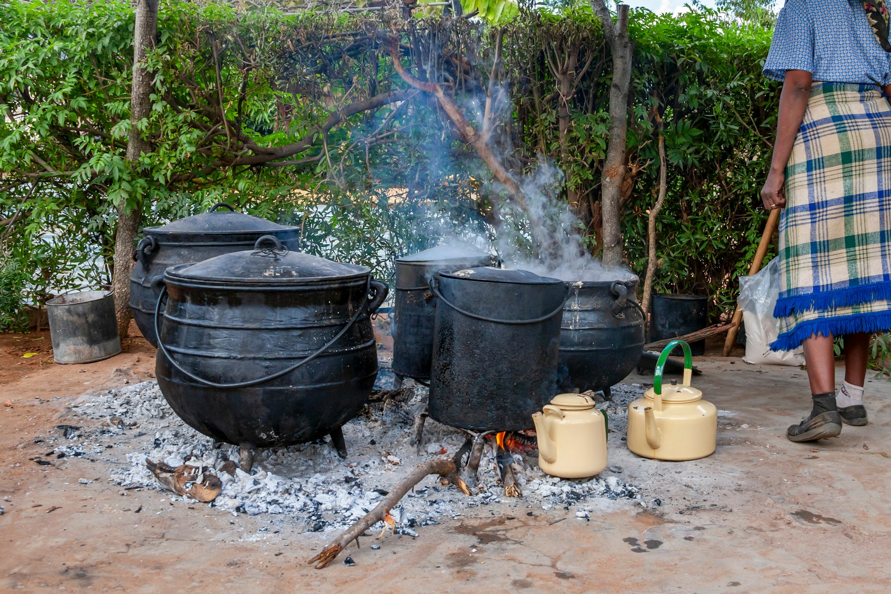 Photo depicts a woman cooking using four black cauldrons over an ashy and wood filled fire