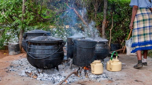 Photo depicts a woman cooking using four black cauldrons over an ashy and wood filled fire