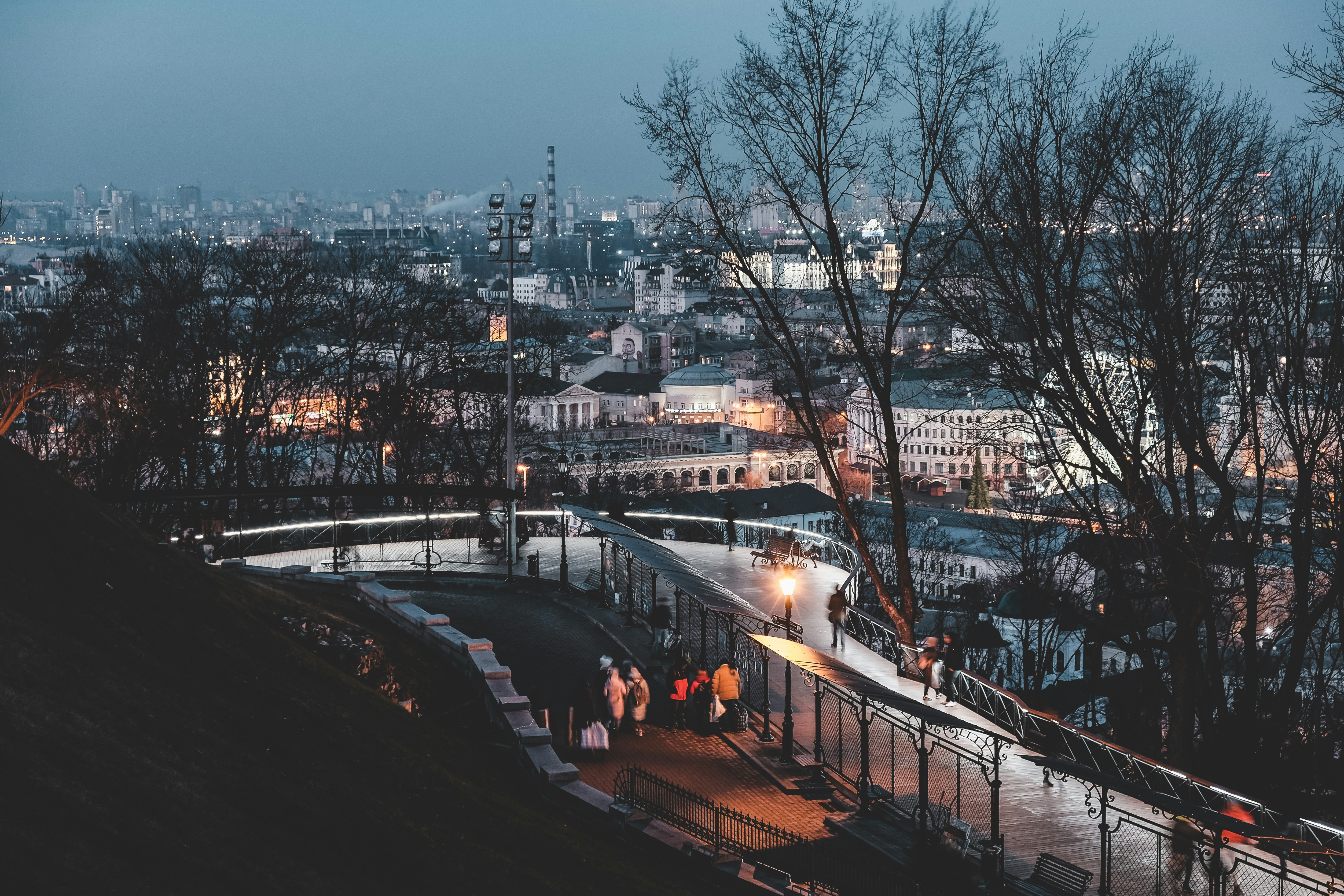 The photo depicts people walking on sidewalk near bare trees during night time in Kyiv, Ukraine.