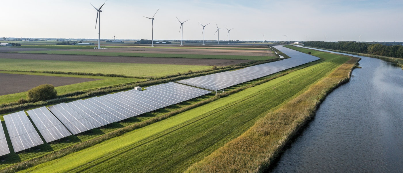 Photo shows a green landscape with a row of modern wind mills and solar panels to produce clean energy. A canal of water flows on the right side of the photo into the horizon,