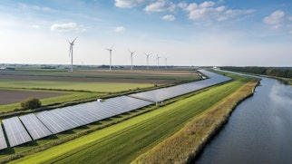 Photo shows a green landscape with a row of modern wind mills and solar panels to produce clean energy. A canal of water flows on the right side of the photo into the horizon,