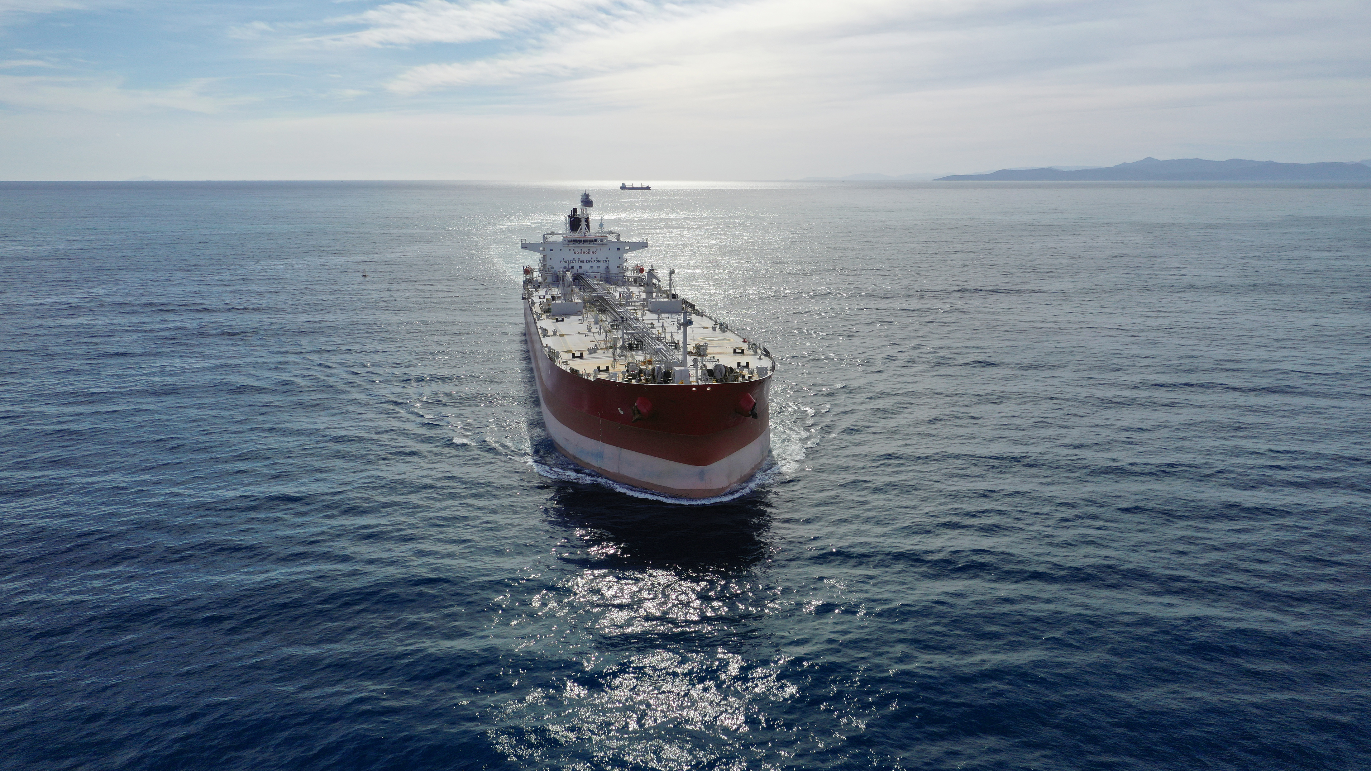 Image depicts a large oil tanker heading face on. it is red with a white stripe and mechanical attributes. The ship is in a large body of water with land in the distance.
