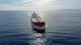 Image depicts a large oil tanker heading face on. it is red with a white stripe and mechanical attributes. The ship is in a large body of water with land in the distance.