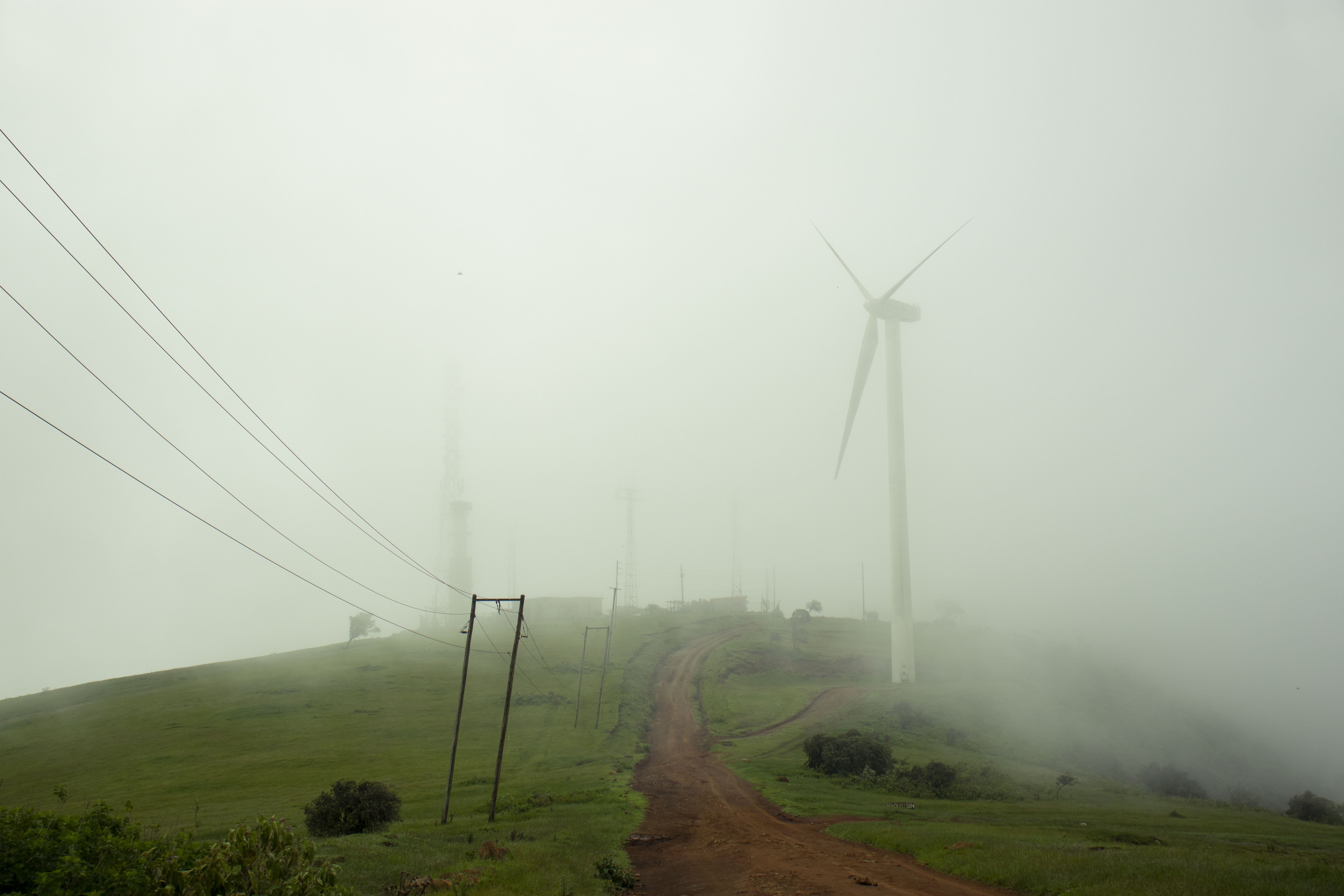 Photo depicts a A dirt road leading up a foggy power pant with wind turbines