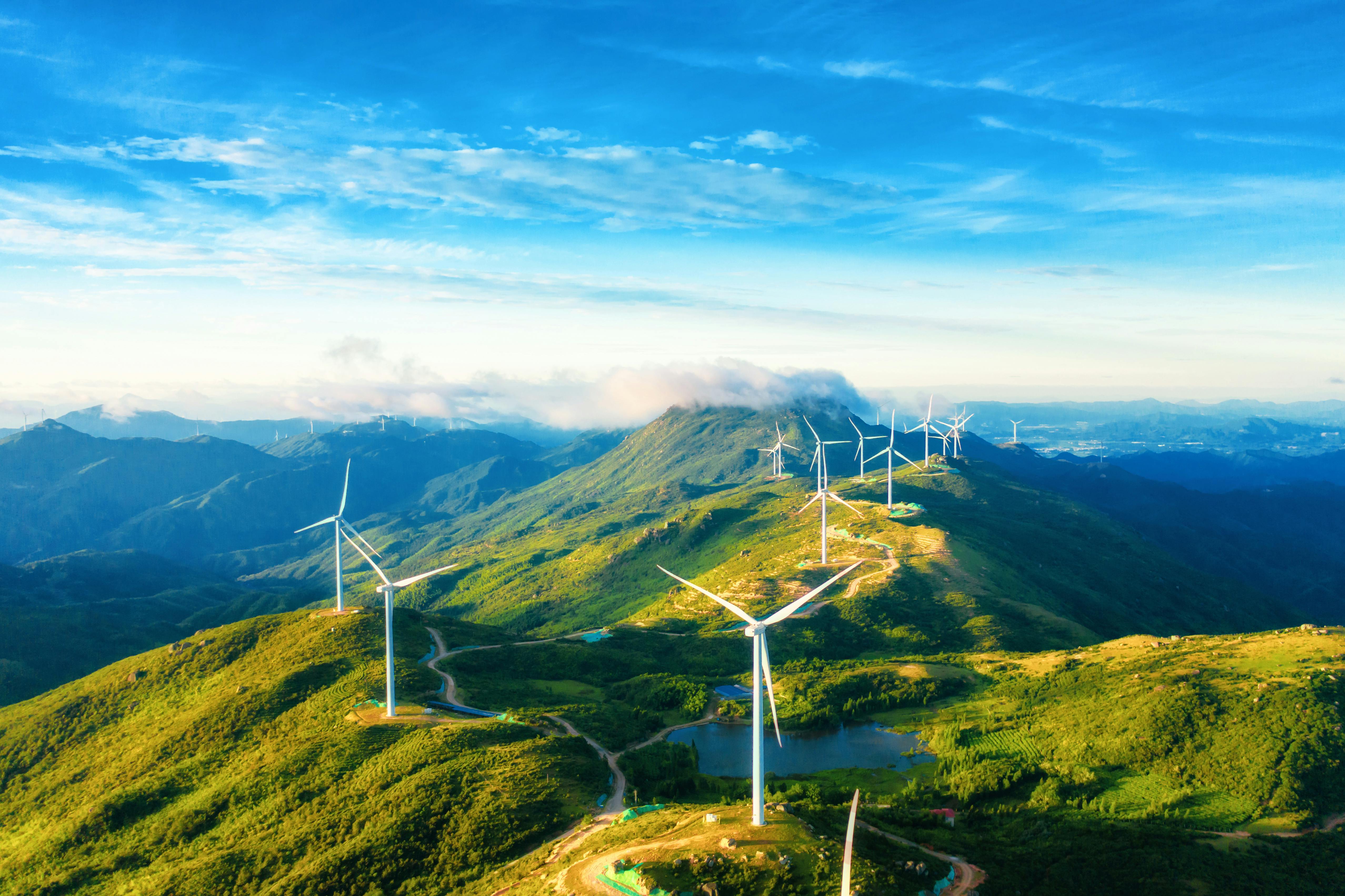 Photo depicts several modern windmills on top of a green mountain range, the windmills get smaller in the distance, the sky is bright blue with white clouds