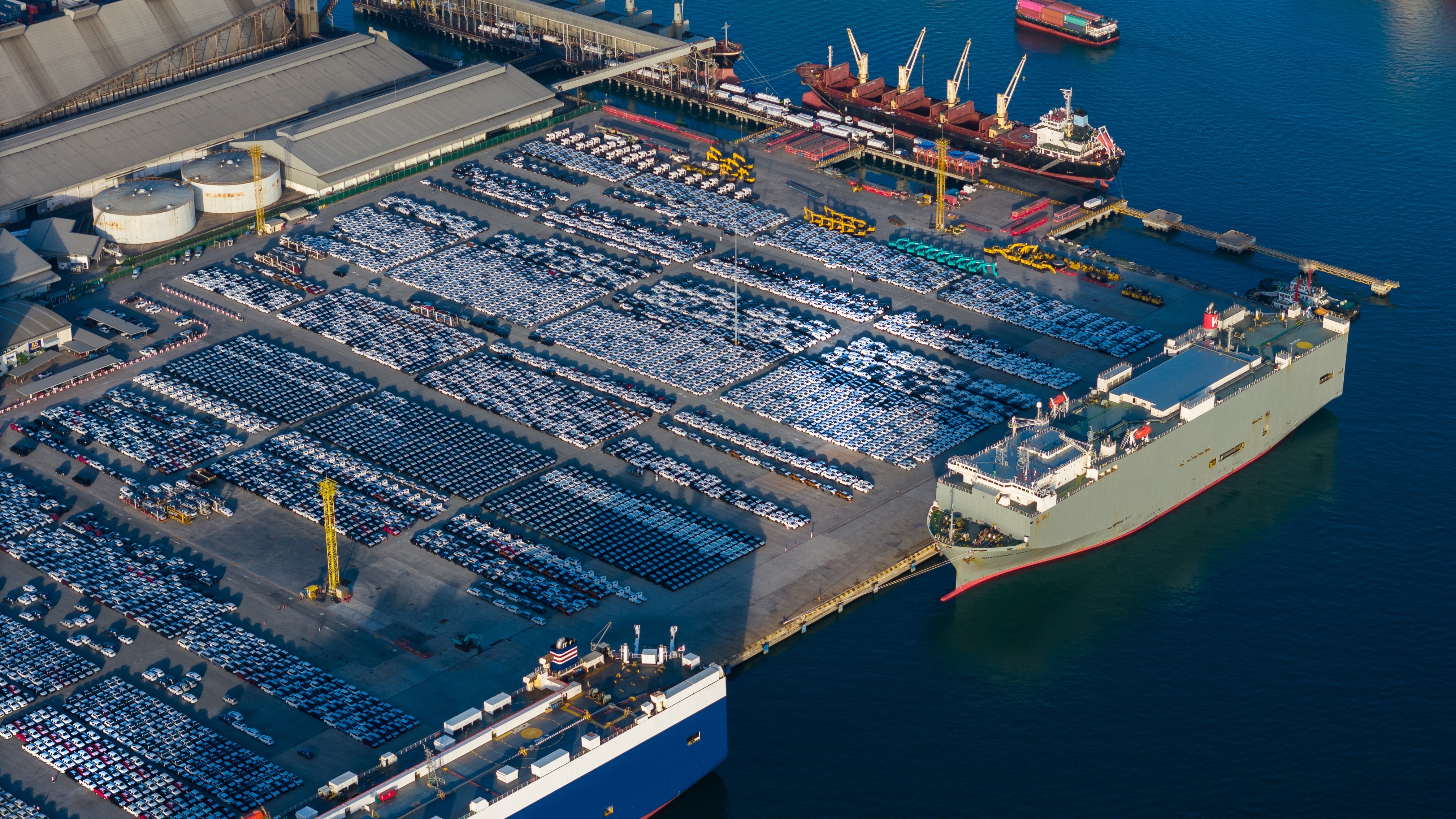 Photo depicts The RoRo (roll-on roll-off) makes a stop at a major industrial port, surrounded by thousands of new vehicles staged for global export, next to busy container transport terminals.