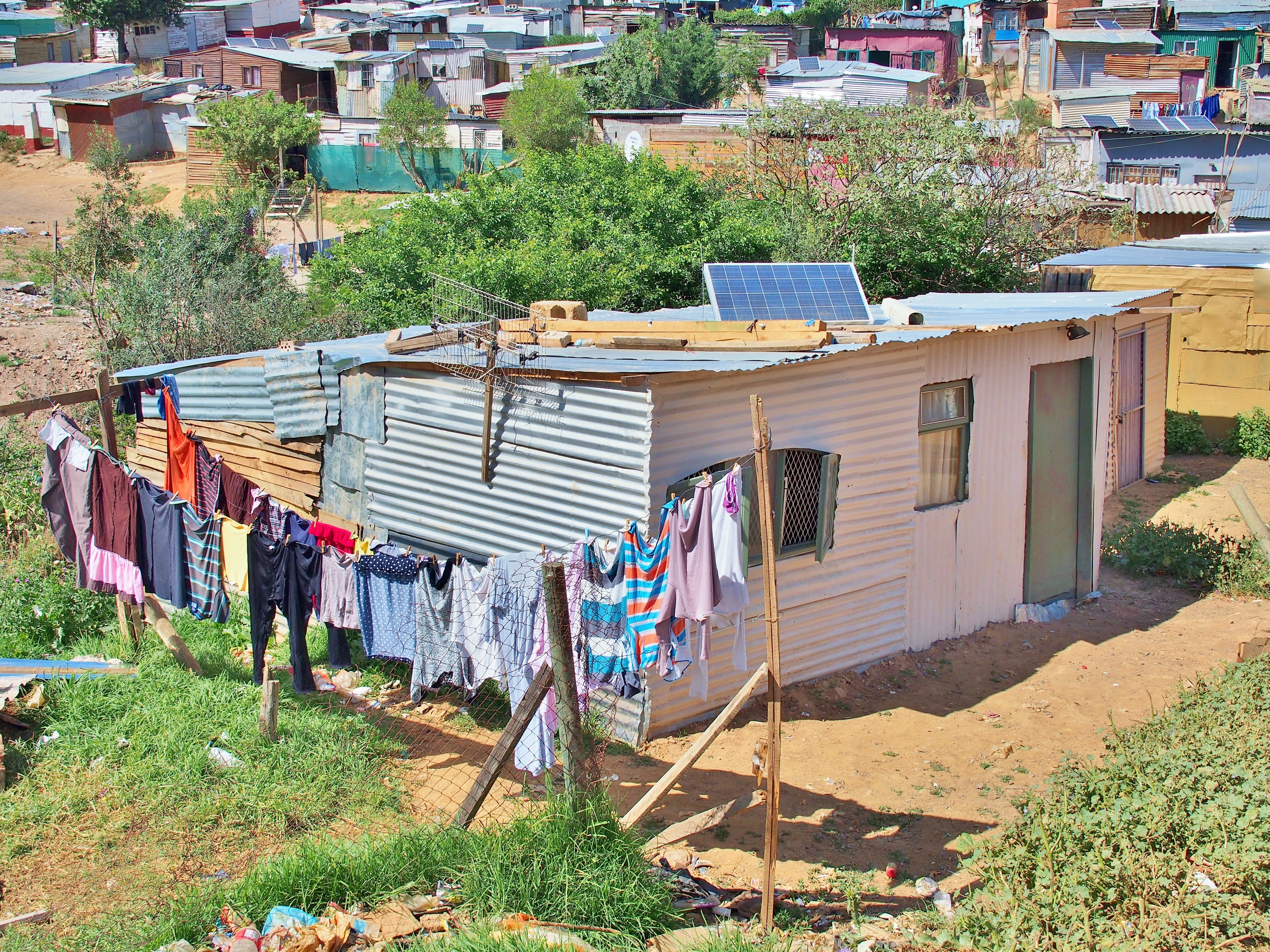 Photo shows a rustic low income shack in india put together by metal panels, a full clothesline is in front of the shack. A solar panel is propped on the roof of the shack. Similar shacks with solar panels are clustered in the distance