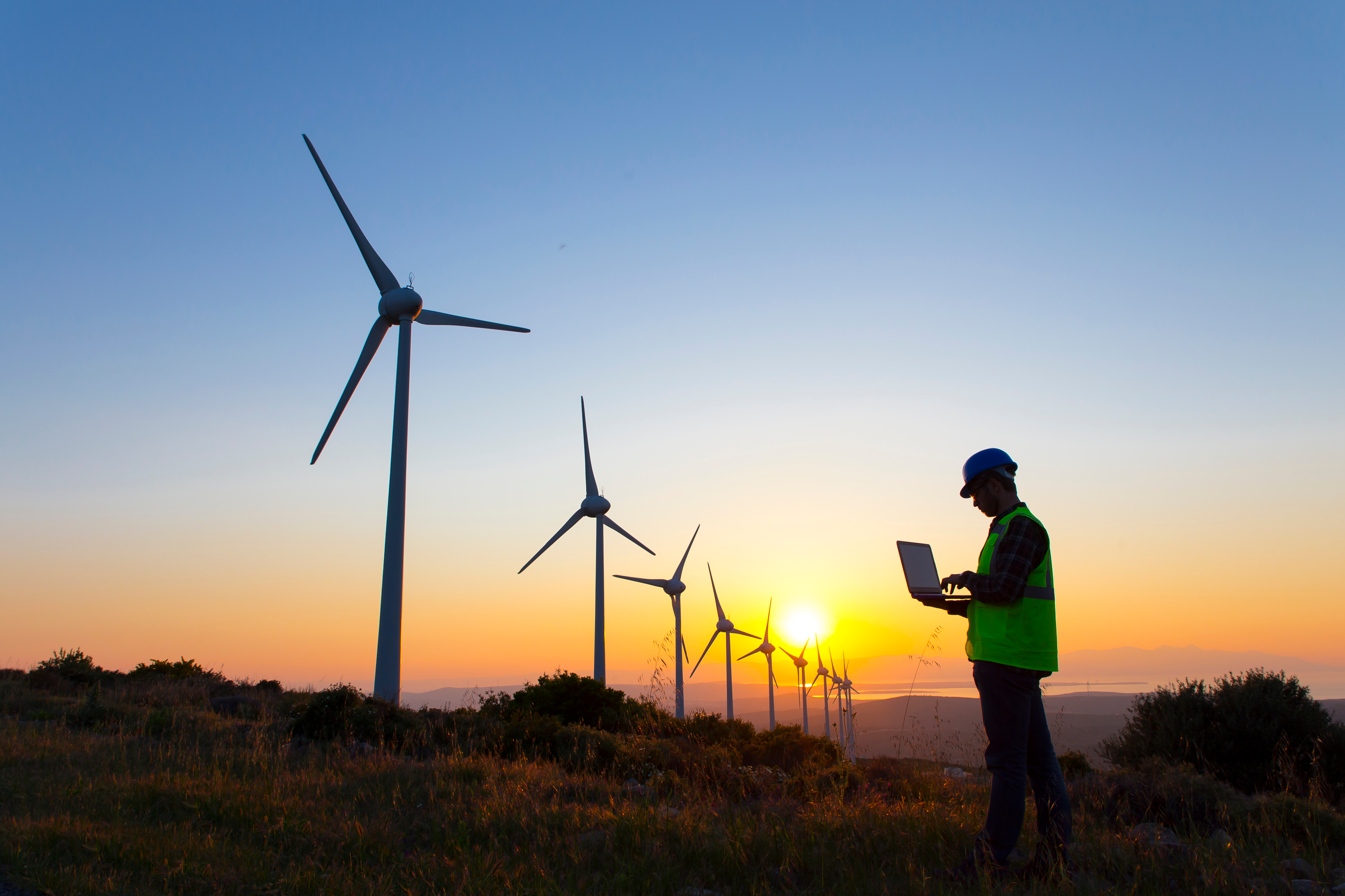 Photo depicts Young maintenance engineer man working in wind turbine farm at sunset
